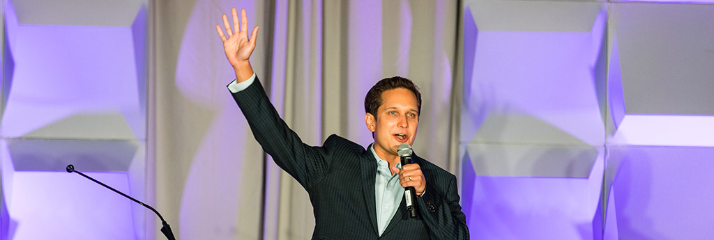 Jason Dorsey speaking on a stage holding a microphone and waving his right arm, purple background lighting